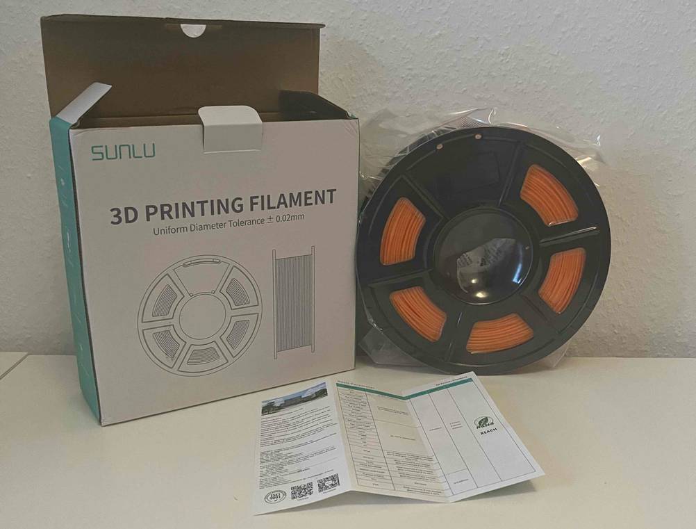 Eine in Zellophan eingeschweißtes 3D-Drucker-Filament mit orangem Drucker-Faden lehnt neben seiner Originalverpackung und Produktbeschreibung an einer weißen Wand.