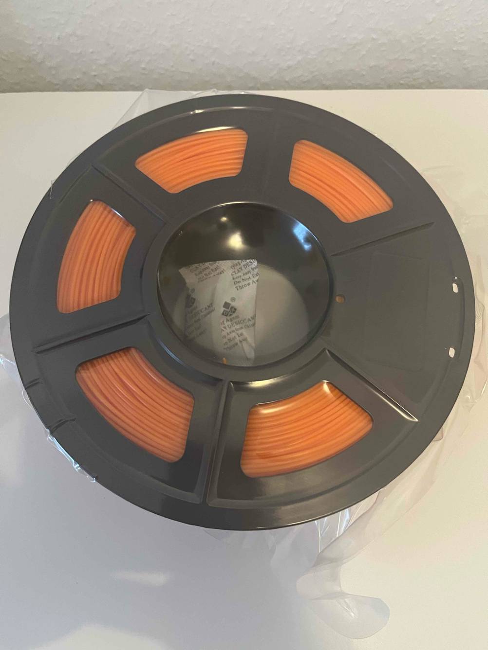 Ansicht von oben auf eine in Zellophan eingeschweißte getestete 3D-Drucker-Filament-Rolle mit orangem Drucker-Faden.