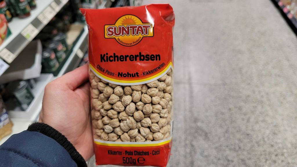 Eine Hand hält eine 500-g-Packung mit getrockneten getesteten Kirchererbsen vor ein Regal im Supermarkt.