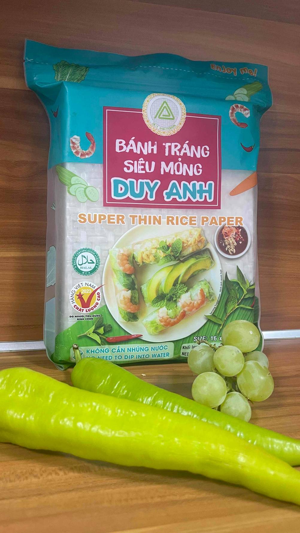 Eine Packung mit dünnem getesteten Reispapier liegt hinter grünen Trauben und zwei Paprika auf einer Holzoberfläche.