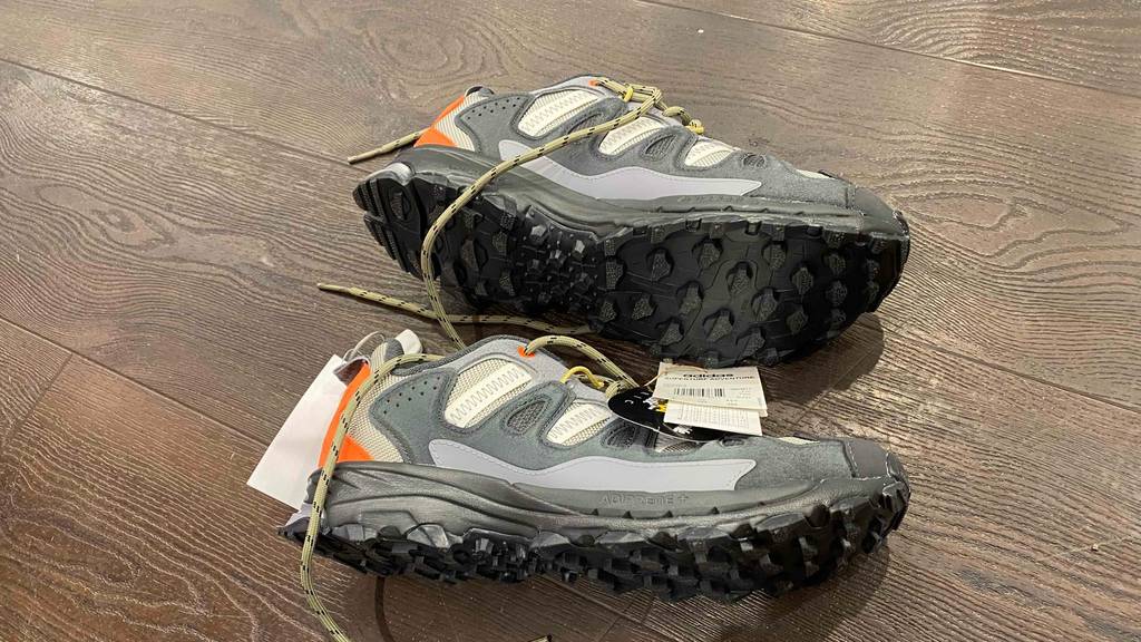 Niedrige beige-graue getestete Herren-Trekkingschuhe liegen mit Fokus auf die orange leuchtende Applikation im Fersenbereich und die ausgeprägte Profilsohle auf der Seite auf einem Holzboden.