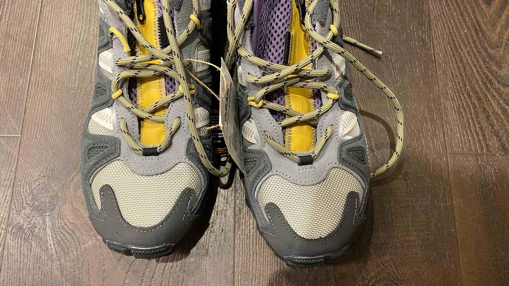 Nahaufnahme der grauen Zehenkappen von niedrigen beige-grauen getesteten Herren-Trekkingschuhe auf braunem Holzboden.