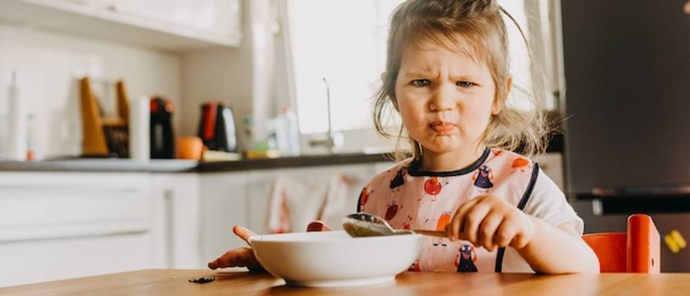 Suppenteller von Kindern getestet: Ein Mädchen sitzt in einer Küche vor einem Suppenteller und hält einen Löffel in der Hand. Sie macht ein unglückliches Gesicht und hat hellbraune Haare.