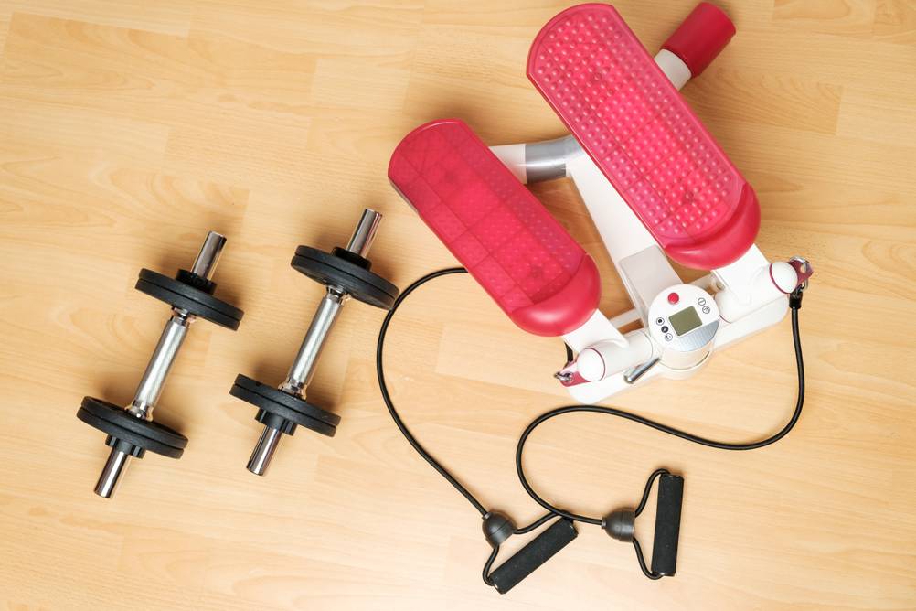 Ein getesteter Swing-Stepper mit pinken Trittflächen, Bändern und Trainingscomputer steht auf dem Boden. Daneben liegen zwei Hanteln. Ansicht von oben.