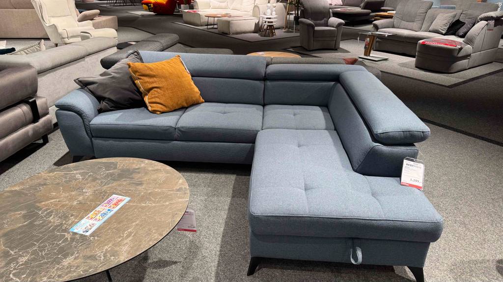 Getestet: Ein Ecksofa mit grauem Stoffbezug und farbigen Kissen steht vor einem ovalen Couchtisch im Ausstellungsbereich eines Möbelhauses.