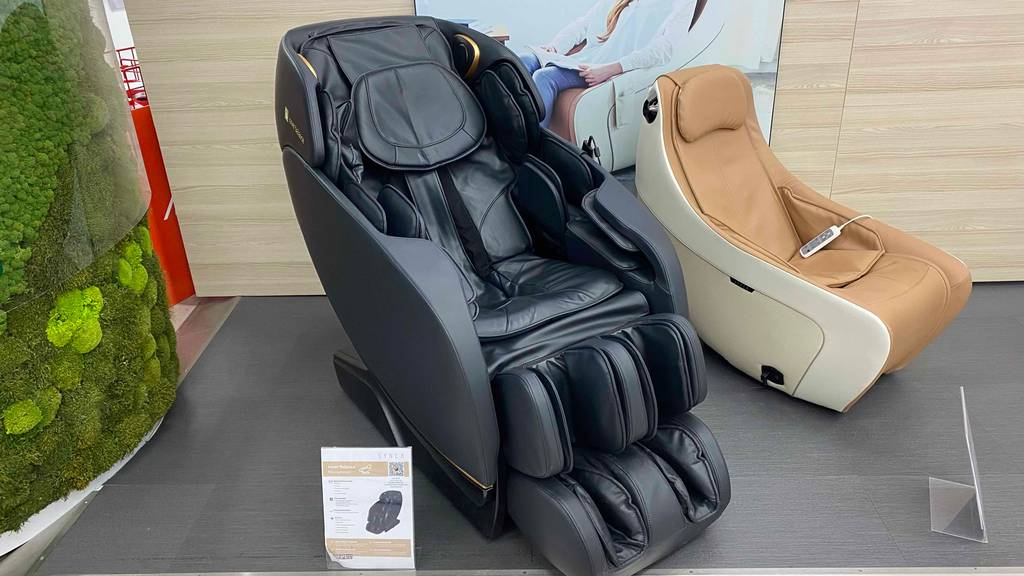 Massagesessel-Test: Schwarzes Modell mit Ganzkörpermassage-Funktionen steht neben einem beigefarbenen Modell in einem Verkaufsbereich.