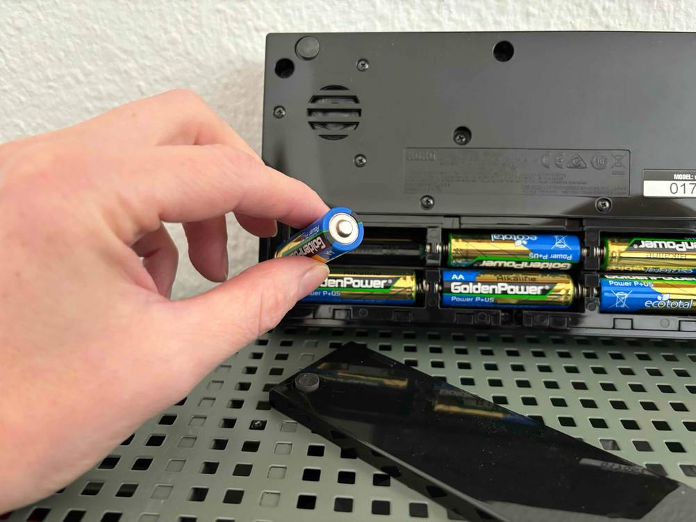 Eine Hand hält eine Batterie, im Hintergrund befindet sich der geöffnete Synthesizer im Test.