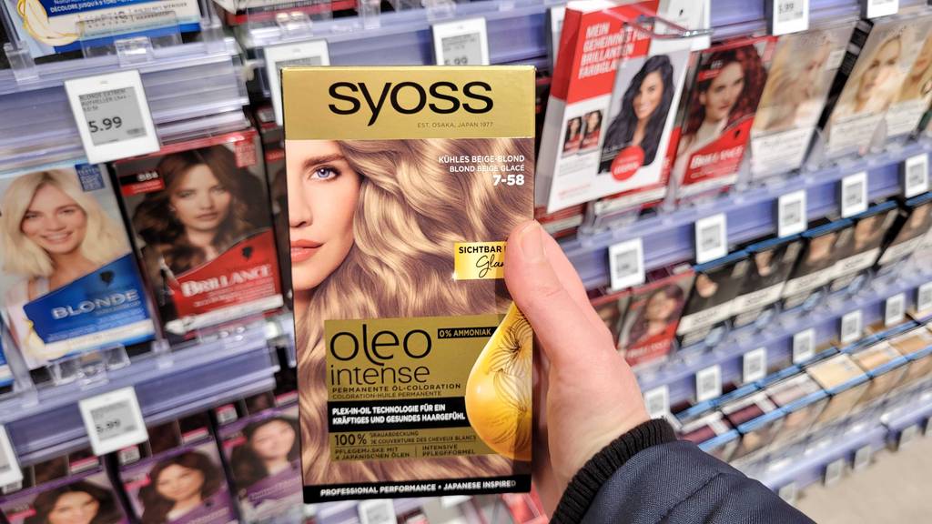 Eine Hand hält eine Packung mit getesteter Haarfarbe von Syoss vor einem Drogeriemarktregal.