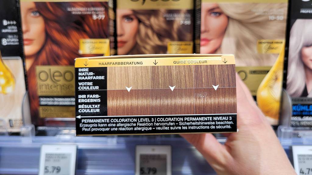 Eine Hand hält eine Packung Haarfarbe von Syoss im Test, inklusive Haarfarbberatung, vor einem Drogeriemarktregal.