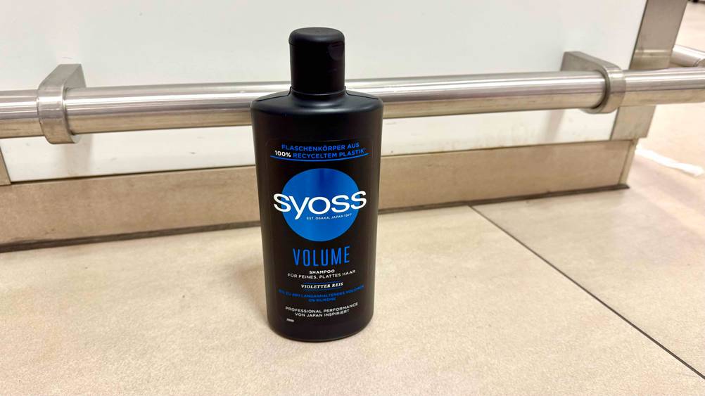 Auf einem beigen Fließenboden steht eine Flasche des getesteten Syoss-Shampoos.