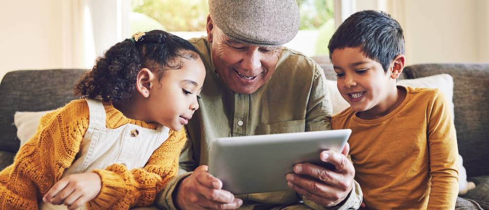 Tablet-Versicherungen-Vergleich: Ein älterer Herr sitzt gemeinsam mit zwei Kindern auf einer Couch, während sie alle auf ein Tablet schauen.