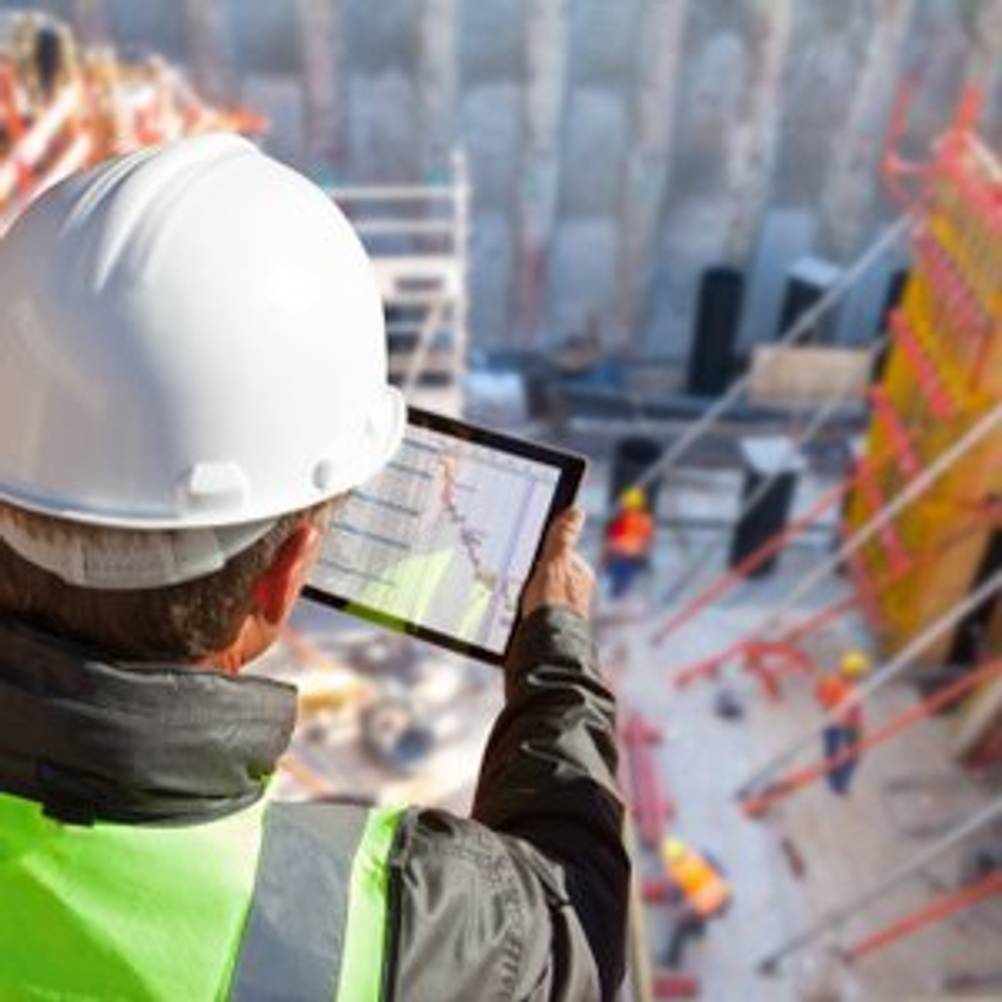 Mann mit Bauhelm und Warnweste, der auf eine Baustelle hinabschaut und dabei ein Tablet in der Hand hält