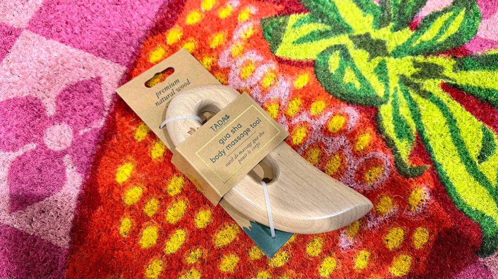 Gua Sha Test: Ein geglättetes Holzstück inklusive Griffmulde liegt in der Originalverpackung auf einem bunten Teppich.