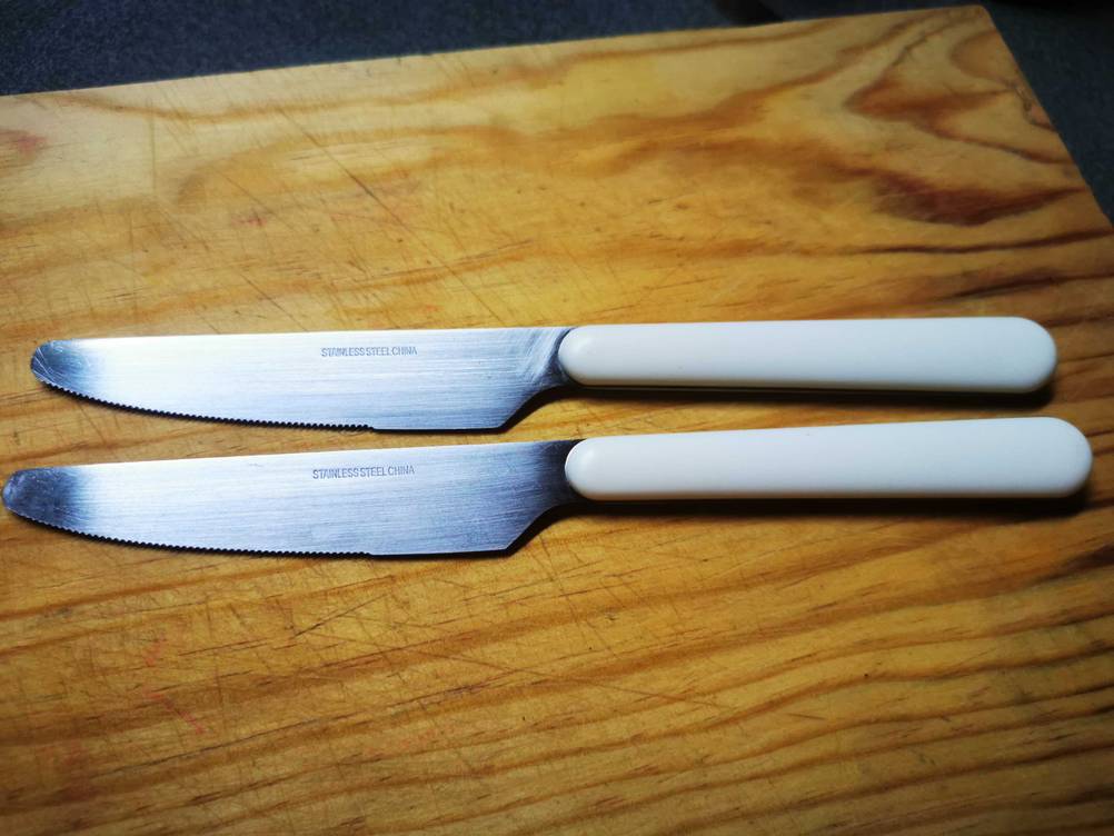 Zwei Tafelmesser im Test mit weißem Griff auf einer Holzfläche.