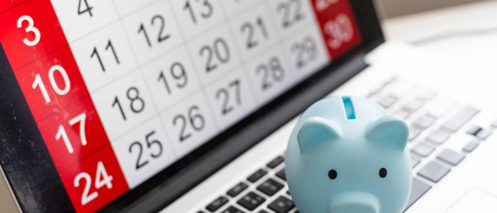 Konten fürs Tagesgeld im Test: Auf einem Laptop, der einen Kalender im Display anzeigt, steht ein hellblaues Sparschwein.