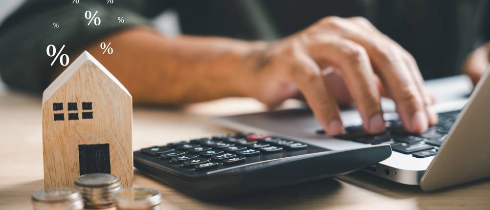 Ein Mann sitzt an einem Laptop, neben ihm liegt ein Taschenrechner, ein Holzhäuschen und mehrere Münzstapel.
