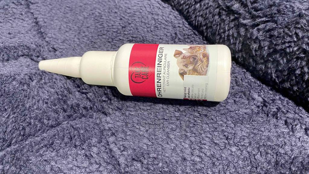 Eine weiße Kunststoffflasche inklusive Pipetten-Verschluss mit Take Care Ohrenreiniger für Hunde im Test liegt auf einem lila Teppich.