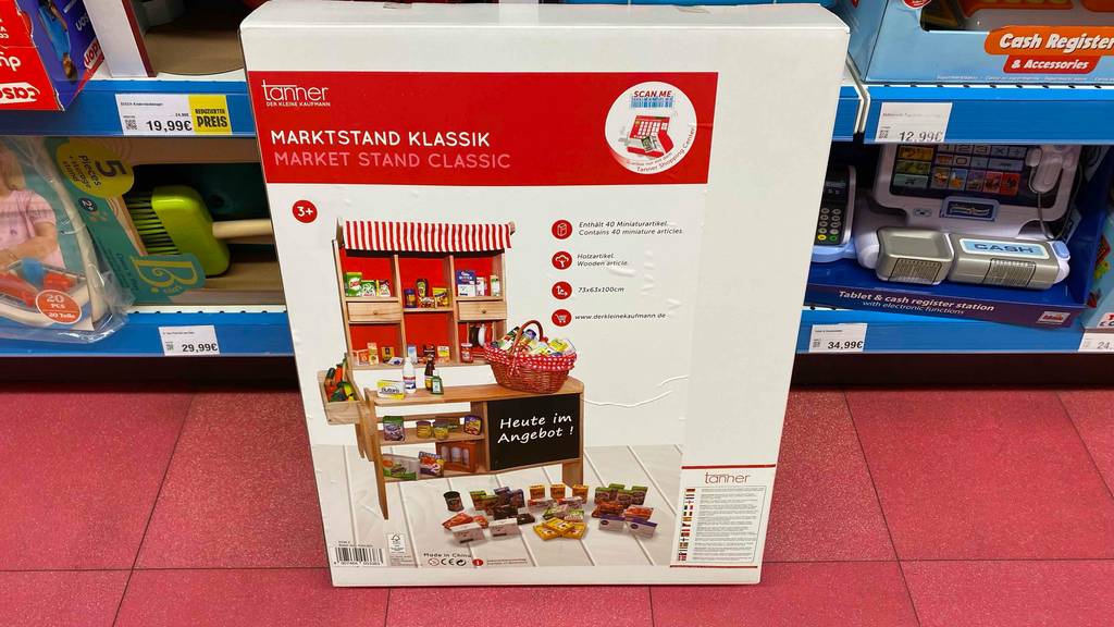 Getestet: Kaufladen Marktstand von Tanner inklusive eine Menge Zubehör steht in seiner Originalverpackung auf einem roten Boden vor einem Verkaufsregal.