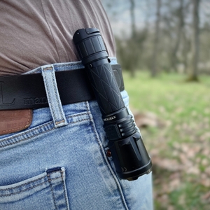 Die Klarus XT21X Pro Taschenlampe ist mit einem Clip sicher an einem Gürtel befestigt. Der Outdoor-Hintergrund deutet auf eine Nutzung im Gelände hin.