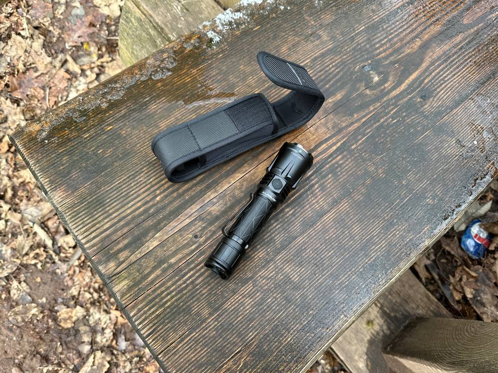 Die Klarus XT21X Pro liegt neben ihrer schwarzen Schutzhülle auf einer Holzbank im Freien. Die Lampe ist ausgepackt und einsatzbereit.