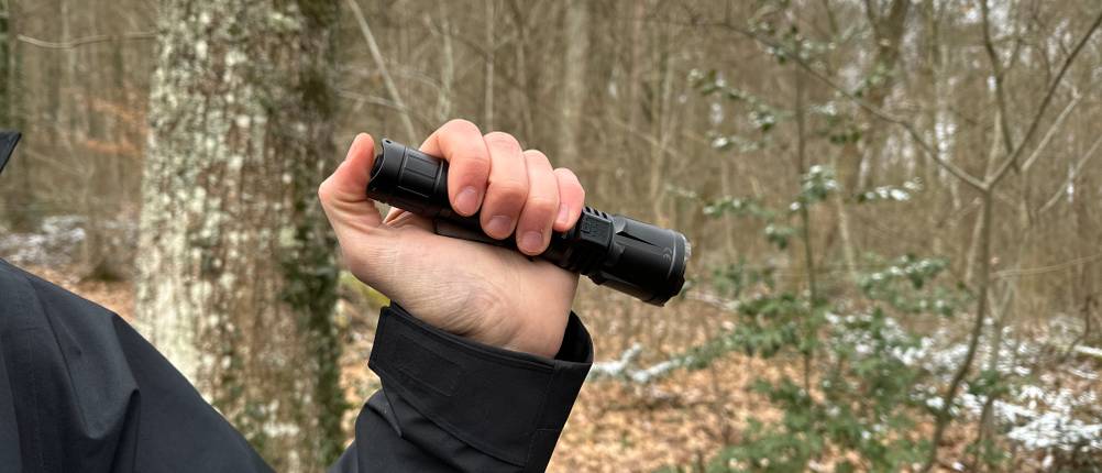 Die Klarus XT21X Pro wird mit einer Hand gehalten, während der Heckschalter bedient wird. Der Outdoor-Hintergrund zeigt eine natürliche Umgebung.