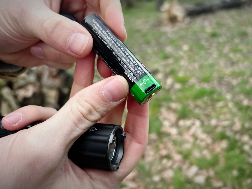 Eine Hand hält den Akku der Nextorch TA30C, auf dem ein USB-C-Ladeanschluss sichtbar ist. Die Taschenlampe wird daneben gehalten.