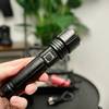 Die Shadowhawk S1476 Taschenlampe wird in einer Hand gehalten. Im Hintergrund ist ihr Zubehör zu sehen.