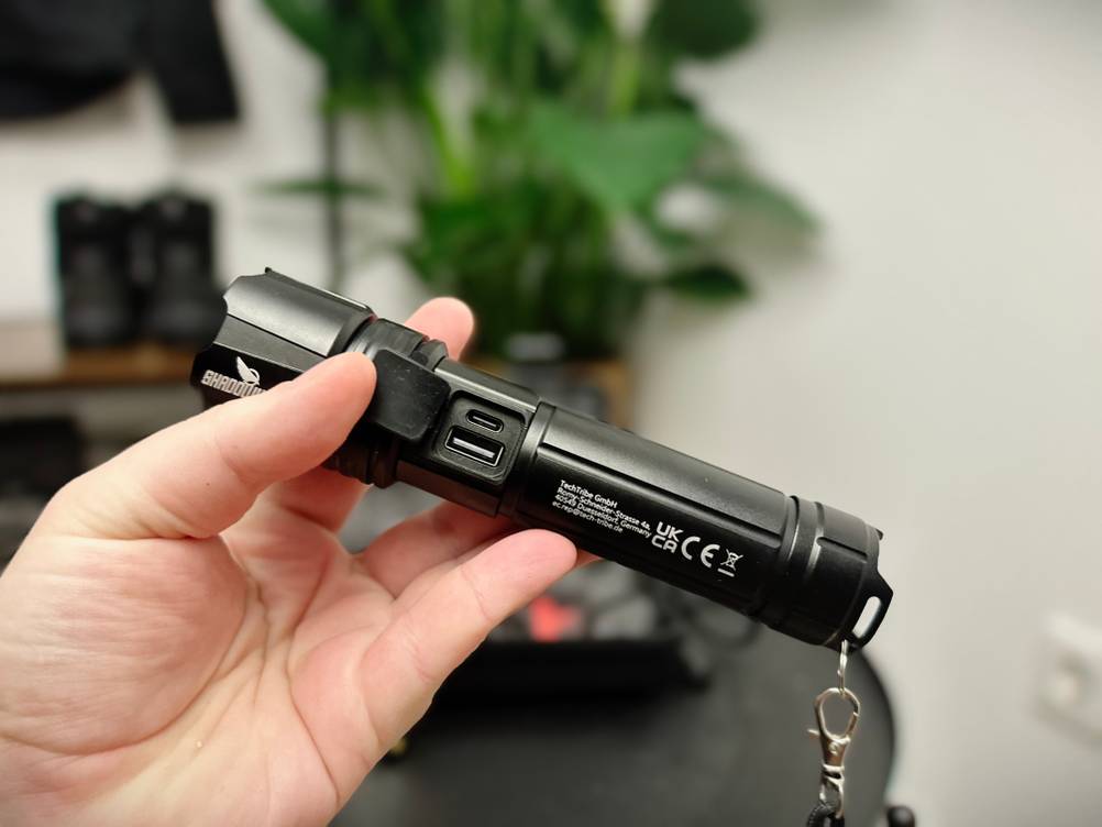 Die Shadowhawk S1476 verfügt über einen abgedeckten USB-C- und USB-A-Anschluss. Eine Hand hält die Taschenlampe, während die Schutzkappe geöffnet ist.