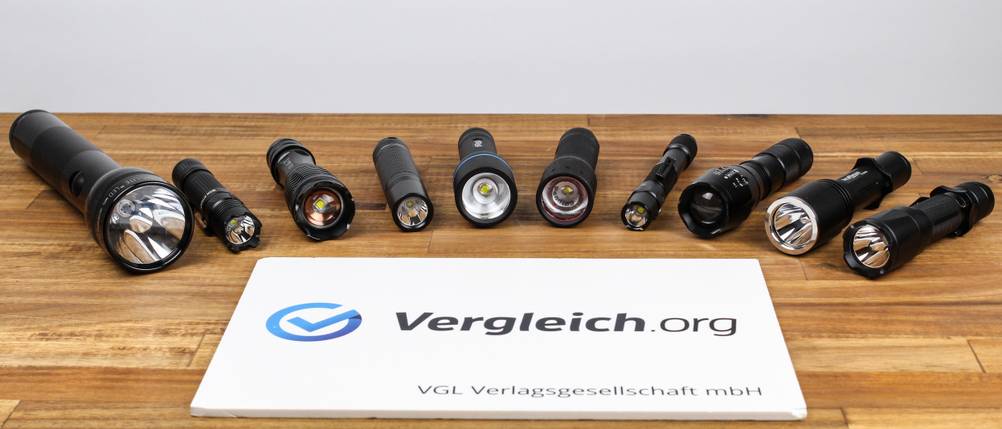 Verschiedene Taschenlampenmodelle stehen in einer Reihe auf einem Holztisch nebeneinander. Im Vordergrund liegt ein Schild mit dem Logo von Vergleich.org.
