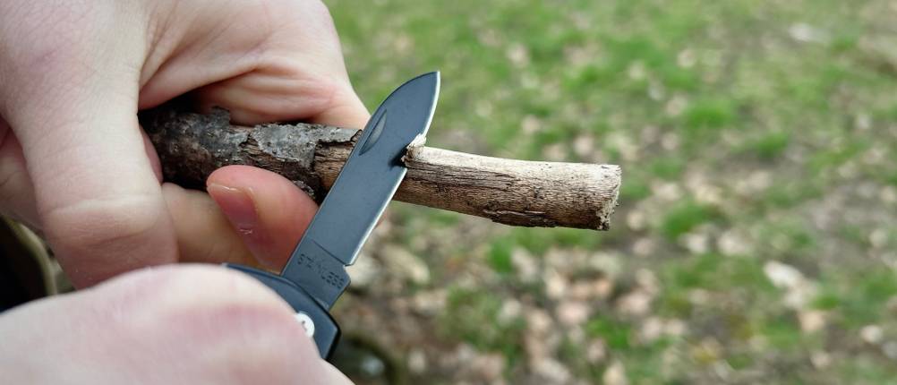 Das Honzin Taschenmesser HJ-011 wird beim Schnitzen eines kleinen Holzasts verwendet. Beide Hände halten das Messer stabil.