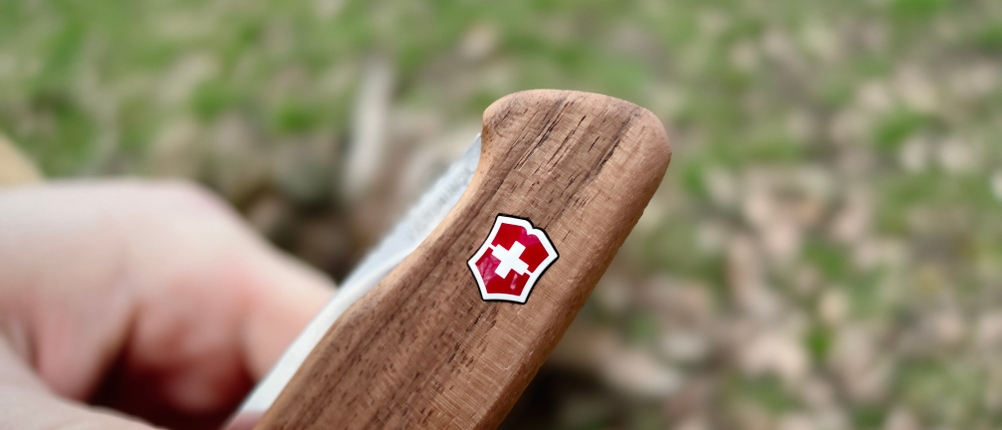 Nahaufnahme des Victorinox-Logos auf dem Holzgriff des Ranger Wood 55. Der Hintergrund zeigt verschwommenen Waldboden.