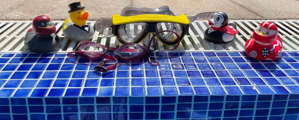 Eine getestete Taucherbrille in Dunkelrot und einer Tauchermaske in Gelb liegen zwischen mehreren Schwimmenten auf einem blau-weißen Beckenrand.