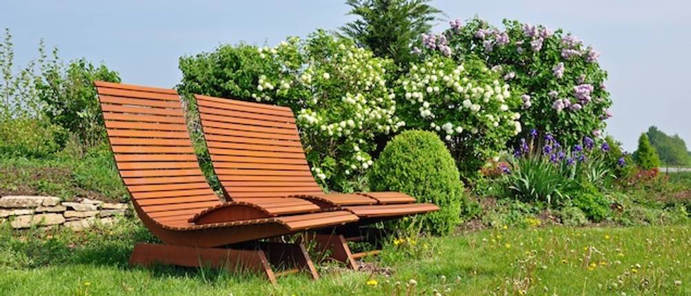Teak-Gartenliegen im Test: Zwei Teak-Gartenliegen stehen nebeneinander auf einer Wiese vor blühenden Hecken.