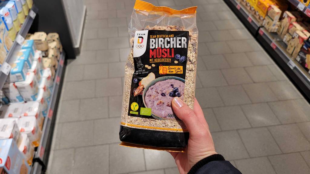 Eine Hand hält eine Packung Bircher-Müsli zum testen über einem Fliesenboden.