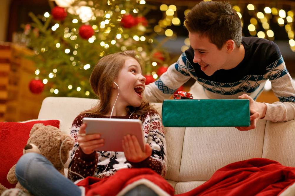 Ein Mädchen hält ein Tablet in den Händchen und dreht sich zur Seite zu einem Jungen. Dabei lacht sie. Im Hintergrund ist ein Weihnachtsbaum zu sehen. Der Junge hält ein Geschenk in der Hand.
