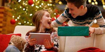 Ein Mädchen hält ein Tablet in den Händchen und dreht sich zur Seite zu einem Jungen. Dabei lacht sie. Im Hintergrund ist ein Weihnachtsbaum zu sehen. Der Junge hält ein Geschenk in der Hand.