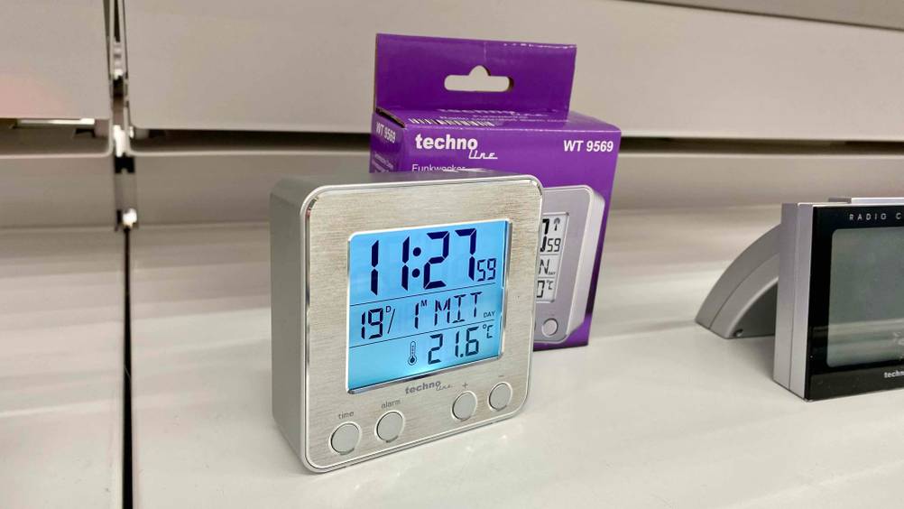 Im Test: Ein Funkwecker mit angeschaltetem Display vor seiner Verpackung in einem Verkaufsregal von schräg vorne.