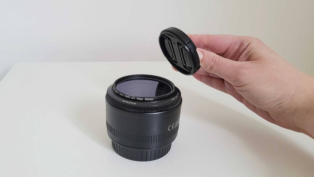 Im Test: Eine Hand hält einen Objektivdeckel über eine auf dem Tisch stehendes Objektiv mit UV-Filter.