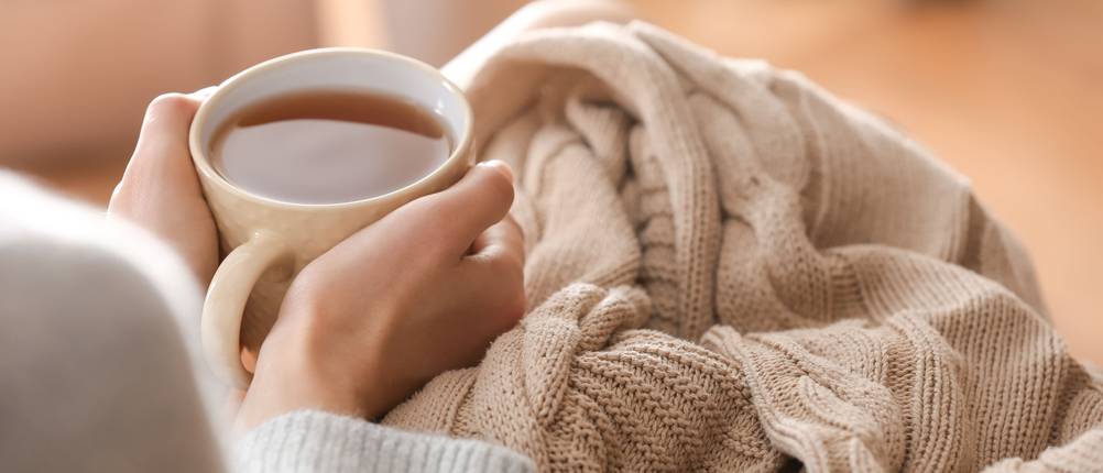 Eine Person ist in einer Wolldecke eingekuschelt und hält eine Tasse Tee in den Händen.