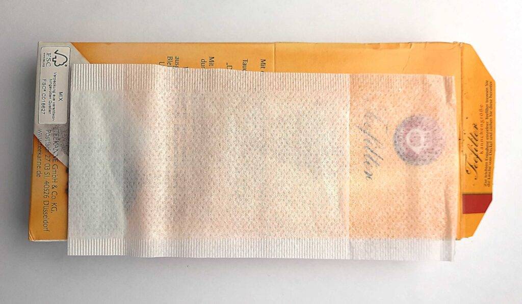 Ein ausgebreiteter transparenter getesteter Teebeutel liegt auf seiner orangen Verpackung.