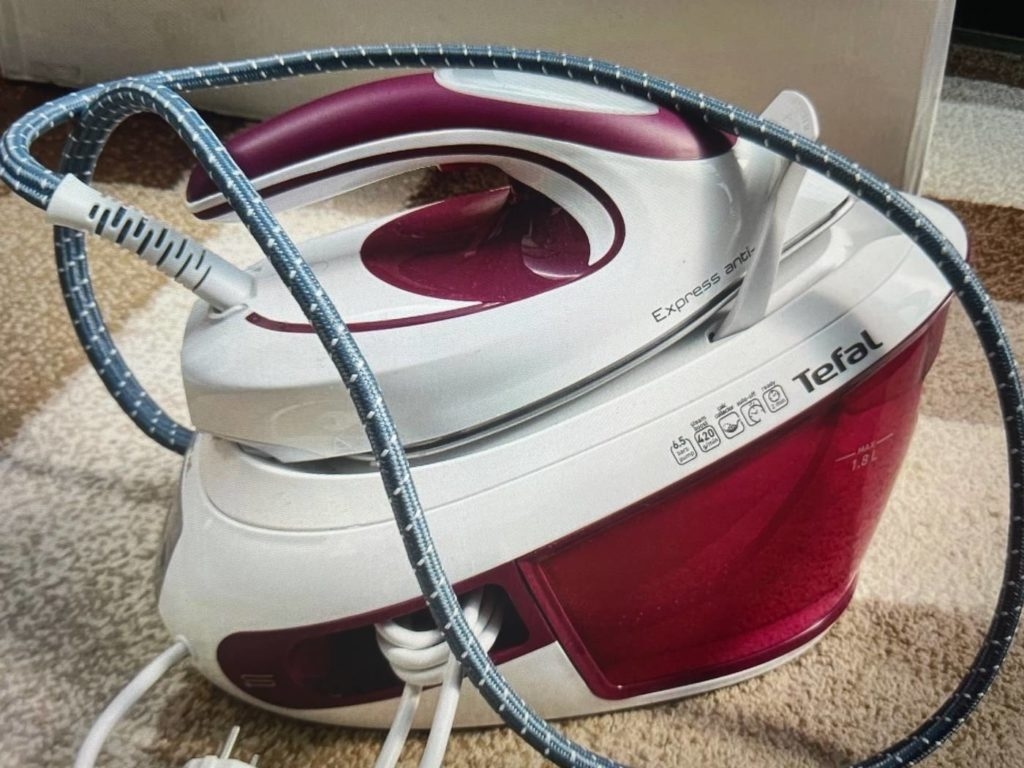 Getestete Dampfbügelstation im weiß-weinroten Design der Marke Tefal steht auf einem beige-braun-karierten Teppich.