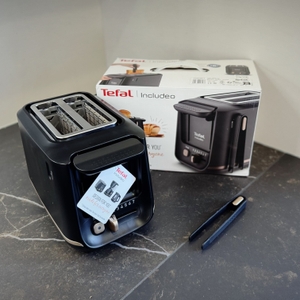 Der getestete Toaster von Tefal steht mit der Verpackung und der Zange auf einer dunklen Arbeitsfläche.