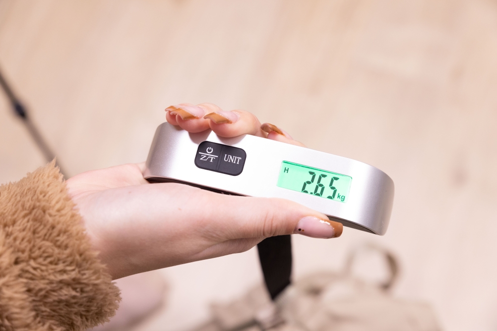 Eine Kofferwaage mit digitaler Anzeige wird in der Hand gehalten.