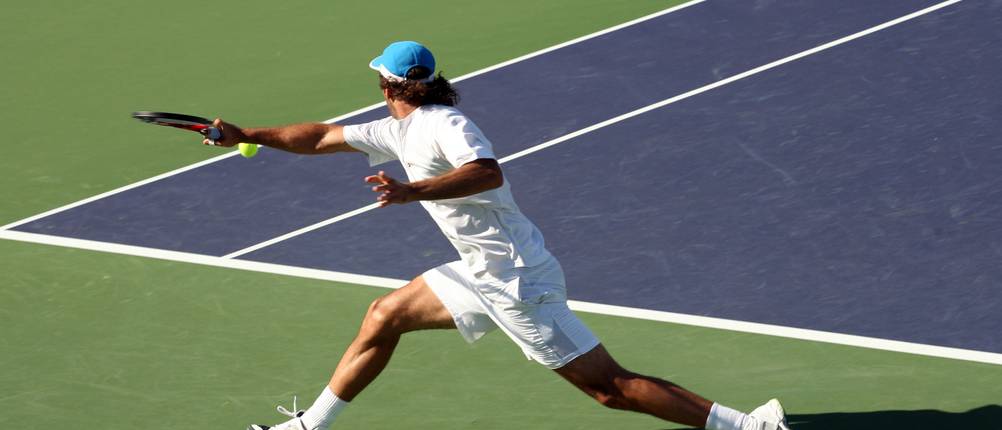 Tennisschuhe-Herren-Test: Zu sehen ist ein Tennisspieler auf einem Outdoor-Hartplatz, während er auf der Rückhandseite in einen Ball rutscht.