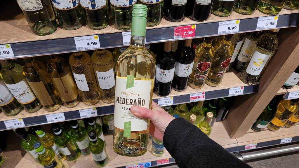 Freixenet-Wein-Test: Eine Hand hält eine Flasche mit Weißwein vor einem Weinregal.