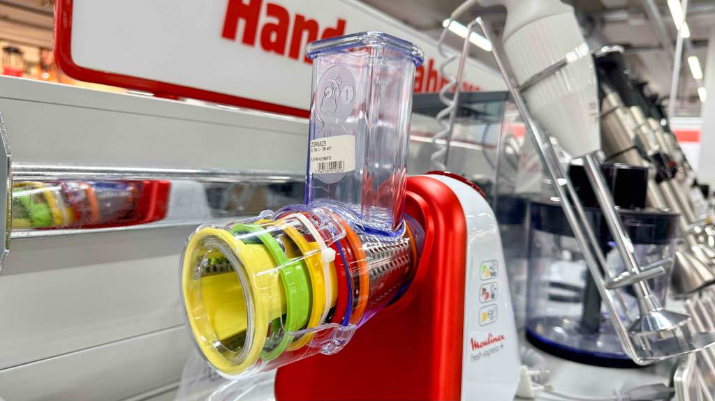 Nahaufnahme der bunt markierten Aufsätze eines getesteten Moulinex-Zerkleinerers, der auf einem Verkaufsregal vor anderen Mixern steht.
