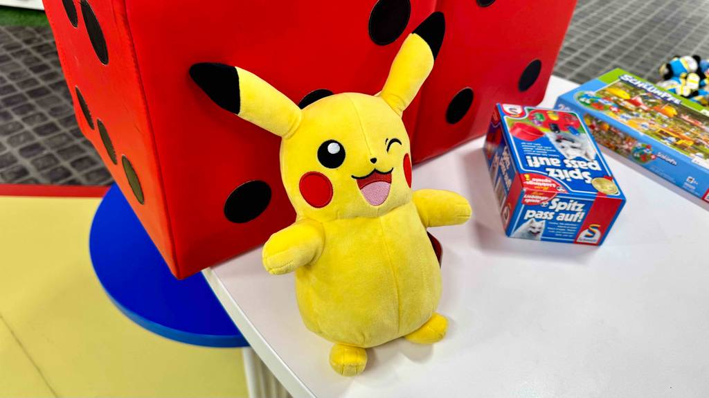 Ein gelbes Pokémon-Kuscheltier im Test lehnt an einem roten Plüsch-Würfel auf einem Ausstellungstisch.