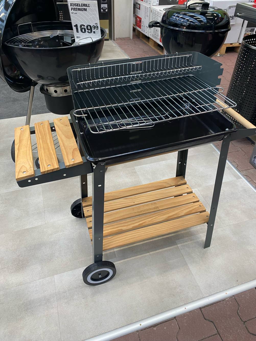 Gesamtansicht eines fahrbaren, getesteten Grillwagens inklusive zwei höhenverstellbarer Grillflächen sowie zwei Holzablagen im unteren und seitlichen Bereich.