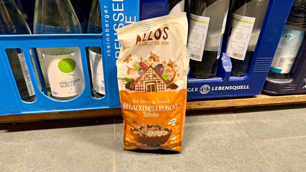 Eine 400-g-Tüte mit Allos Müsli im Test steht vor blauen Getränkekästen.