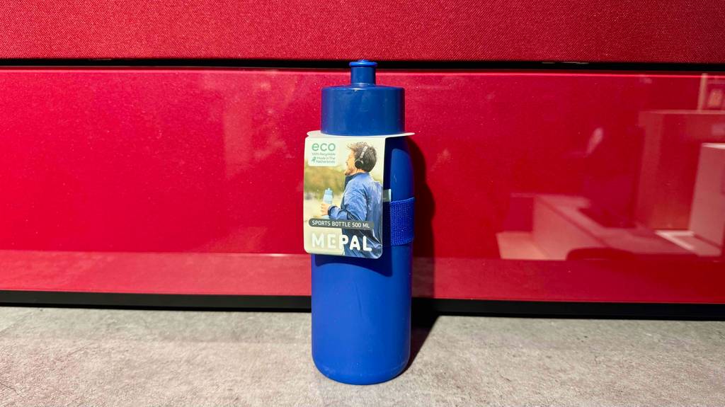 Eine blaue Mepal-Trinkflasche im Test steht auf einem Boden vor einer roten Regalwand.